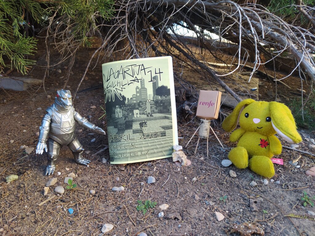 Fotografía de lanzamiento del decimocuarto número.
se puede ver 4 objetos, de izquierda a derecha se ve un muñeco de Mechagodzilla, el fanzine de este mes, de color amarillo, un muñeco que se compone de palillos como patas y brazos, un corcho de vino como cuerpo y un cartón de Renfe como cabeza, y, finalmente un osito de peluche amarillo.
El fondo se pueden ver arboles, en el suelo hay tierra y un puñado de arboles