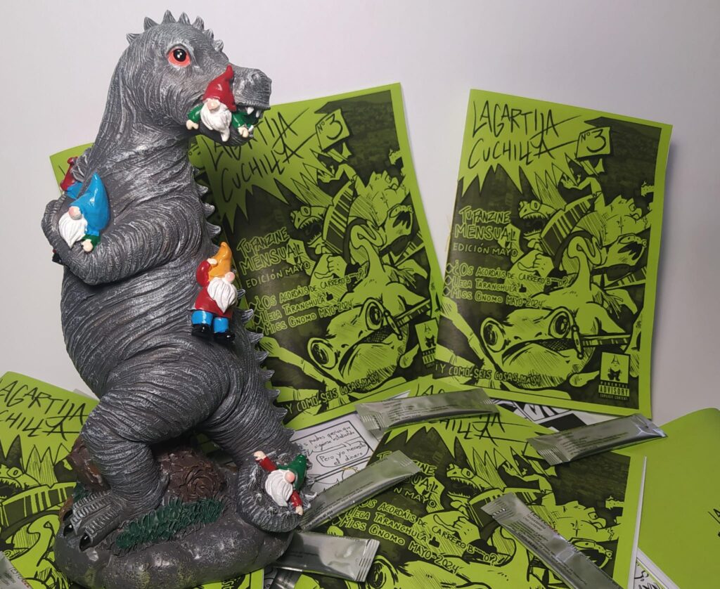 Fotografía del lanzamiento del tercer número, a la izquierda de la foto hay una estatua de unos gnomos siendo atacados por un dinosaurio
a la derecha aparecen los fanzines del tercer número, de color verde. 