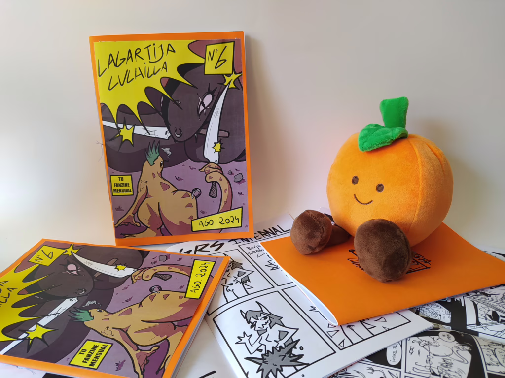 Fotografía de lanzamiento del sexto número, en él, aparece un muñeco naranja sentado encima de un puñado de fanzines de este número a la derecha. a la izquierda se puede ver la portada frontalmente.