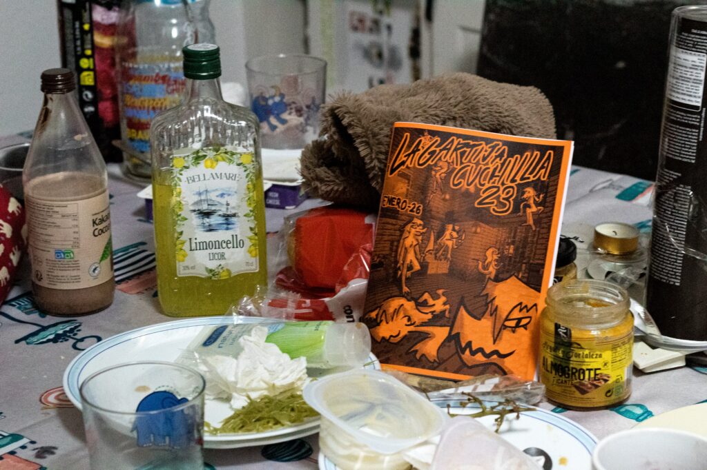 Fotografía del vigecimotercer número. Aparece un comedor muy desordenado lleno de cosas, entre ellos platos sucios, un lixor, almogrote. en la derecha se encuentra el fanzine de este mes, de color naranja.